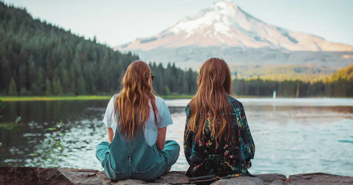 Twee vriendinnen zitten naast een meer met bergen op de achtergrond
